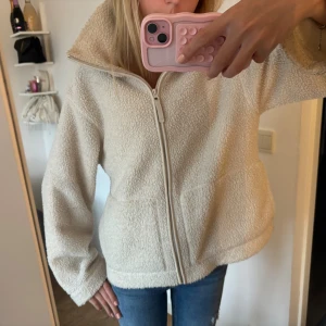 Beige teddyjacka med dragkedja - Mysig beige teddyjacka med hög krage och hel dragkedja framtill. Jackan har en loose passform och är tillverkad i fluffigt fleece-material som ger en chill och trendig vibe. Perfekt för lager-på-lager och att hålla sig varm under kyliga dagar. Lite smink vid kragen.