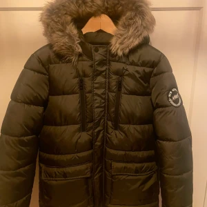 Svart pufferjacka med fuskpäls - Svart pufferjacka från MONITOr med stor huva kantad av fuskpäls. Jackan har två vertikala bröstfickor med dragkedja och ett emblem på ärmen. Perfekt för kalla dagar och riktigt snygg till vintern.