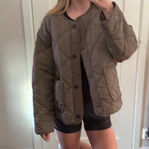 Quiltad khakigrön jacka med knappar - Säljer en oversized quiltad jacka i brun/grön nyans med stora fickor och knäppning framtill. Jackan har rund halsringning och lång ärm, perfekt för lager-på-lager. Snygg och trendig modell som passar till många outfits.