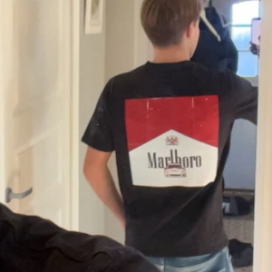 Svart Marlboro t-shirt med tryck - Cool svart t-shirt med stort Marlboro-tryck i rött och vitt på ryggen. Klassisk passform och korta ärmar, perfekt för dig som gillar retro och statement-plagg. Materialet känns som mjuk bomull och trycket är riktigt tydligt.