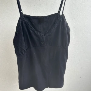 Svart linnetopp med spets och knappar - Svart linnetopp med justerbara smala axelband, v-ringad hals med spetskant och två knappar framtill. Det är i ett linne material.