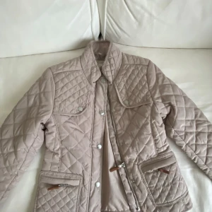 Beige quiltad jacka från Zara Girls - Quiltad beige jacka från Zara Girls med snygga detaljer som tryckknappar, dragkedjor och bruna läderdetaljer på fickorna. Jackan har hög krage och långärmad passform, perfekt för kyligare dagar. Stilren och enkel att matcha med allt. Barn 6/7 (122 cm)