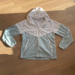 Nike vindjacka med huva, vit och ljusblå - Nike vindjacka i vitt och ljusblått med huva och hel dragkedja framtill. Jackan har två sidofickor med dragkedja och meshfoder på insidan. Swoosh-logga på bröstet. Perfekt för dig som gillar sportig och clean stil.