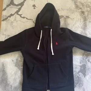 Svart hoodie från Polo Ralph Lauren - Svart hoodie från Polo Ralph Lauren med vit snörning och dragkedja. Klassisk röd logga broderad på bröstet och stora fickor framtill. Perfekt för dig som gillar stilren streetwear med premiumkänsla.