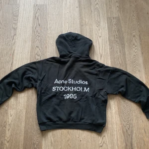 Svart hoodie Acne Studios Stockholm - Svart hoodie från Acne Studios med vit text på ryggen: 'Acne Studios STOCKHOLM 1996'. Klassisk passform med huva. Perfekt för dig som gillar stilrena streetwear-plagg med tydlig logga.