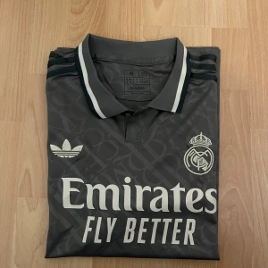 Grå Real Madrid fotbollströja Adidas - Snygg grå Real Madrid fotbollströja från Adidas med korta ärmar och klassisk krage. Tröjan har ett subtilt mönster med RM och CFC över hela tyget, vita detaljer på krage och ärmslut, samt broderade loggor för Adidas och Real Madrid. Materialet är lätt och andas, perfekt för match eller träning.