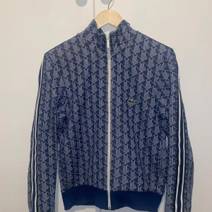 Lacoste Monogram - Säljer en blå och vit mönstrad ziptröja från Lacoste med klassisk krokodillogga på bröstet. Tröjan har hel dragkedja, ståkrage och vita ränder längs ärmarna. Regular fit och perfekt för en sportig look. Skulle säga att tröjan passar Xs mycket.