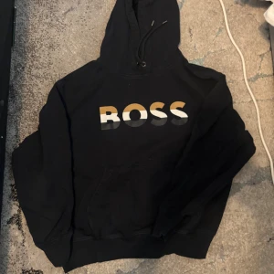 Svart BOSS hoodie med tryck - Svart hoodie från BOSS med stort tryck i vitt, guld och grått på bröstet. Klassisk huva med snörning och magficka framtill. Perfekt för dig som vill ha en stilren och bekväm tröja med statement.