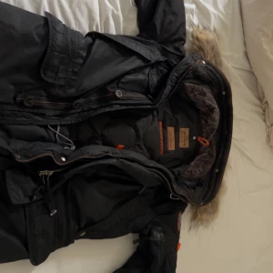 Parajumper ParajumperParajumpers Kodiak Masterpiece Series - Storlek S Vinterjacka - Säljer en svart Parajumpers vinterjacka med pälskantad huva och quiltad insida. Jackan har flera fickor, dragkedja och knappar framtill samt detaljer i orange. Perfekt för kalla dagar och riktigt snygg streetstil.