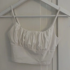 Vit croppad linnetopp med rynk - Säljer en vit croppad linnetopp med smala axelband och snygga rynkdetaljer över bysten. Toppen har en enkel och stilren design som passar perfekt till sommaren. Materialet känns mjukt och lätt mot huden. Aldrig använd