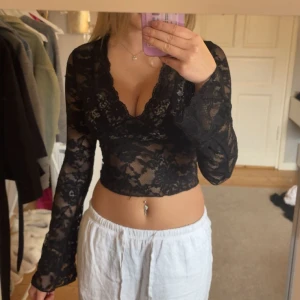 Svart spetsig croppad långärmad topp - Säljer en svart croppad topp i genombruten spets med v-ringning och långa ärmar. Toppen har ett blommigt spetsmönster och är figurnära med snygg passform. Perfekt för att lyfta en outfit med lite extra edge.