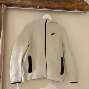 Vit Nike hoodiejacka - Nike hoodiejacka i vitt med svarta dragkedjor och detaljer. Jackan har huva, två fickor med dragkedja och svart Nike-logga på bröstet. Köpt på barnavdelningen men har en annan nike tech från damavdelningen som e i strl S så denna jag nu säljer är jätte stor, de är därför jag säljer den, den är använd ett fåtal gånger.