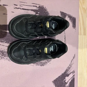 Svarta adidas sneakers strl 21 (barn) - Säljer ett par svarta adidas sneakers med klassiska tre ränder på sidan och gulddetaljer på plösen. Skorna har snörning och en rund tå, tillverkade i syntetmaterial med platt sula. Perfekt för dig som gillar sportig och clean stil.