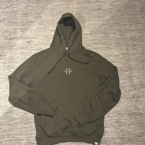 Olivgrön hoodie från Les Deux storlek L - Snygg olivgrön hoodie från Les Deux med broderad logga på bröstet. Tröjan har huva med snörning, ribbade muddar och känguruficka. Perfekt för dig som gillar stilrena och bekväma plagg med en clean look. 