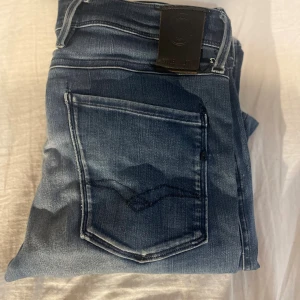 Replay Anbass jeans blå - Snygga Replay Anbass jeans i blå tvätt med slitningar och lätt urtvättad look. Klassisk femficksmodell med raka ben och normal passform. Jeansen har detaljerade sömmar och Replay-logga på fickan. Perfekta för dig som gillar en avslappnad men stilren vibe.