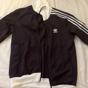 Adidas track jacket med vita detaljer - Mörklila/svart Adidas track jacket med klassiska vita ränder längs ärmarna, vit krage och muddar. Jackan har dragkedja framtill, två fickor med dragkedja och Adidas logga på bröstet. Tillverkad i polyester för en sportig och bekväm känsla.