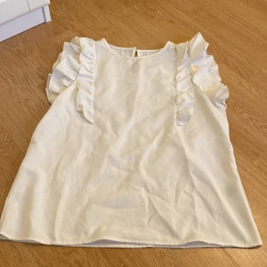 Vit blus med volangdetaljer - Säljer en söt vit blus med volanger längs axlarna och ärmlös design. Blusen har en rund halsringning och en liten pärlknapp i nacken som fin detalj. Materialet känns mjukt och har en lätt glans, perfekt för en fräsch look.
