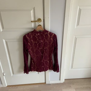 Vinröd spetsblus från Dry Lake - Supersnygg vinröd blus från Dry Lake i spets med blommigt mönster. Hög halsringning, långa ärmar med volangdetalj vid ärmslut och figurnära passform. Perfekt för dig som vill sticka ut med en elegant och trendig look.
