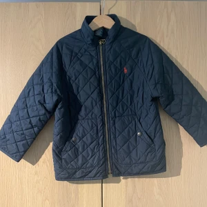 Quiltad jacka från Polo Ralph Lauren - Mörkblå quiltad jacka från Polo Ralph Lauren med dragkedja framtill, två fickor med tryckknappar och klassisk röd logga på bröstet. Jackan är tillverkad i polyester och har en stilren, tidlös look som passar perfekt till höst och vår.