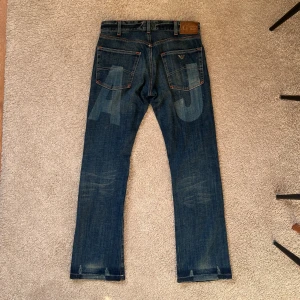 Armani Jeans Bootcut - Snygga blå jeans från Armani Jeans med raka ben och klassisk femficksdesign. Jeansen har tydliga slitningar, orangea sömmar och stora AJ-tryck på bakfickorna. Tillverkade i robust denim med detaljerade nitar och logotyp på fickan.