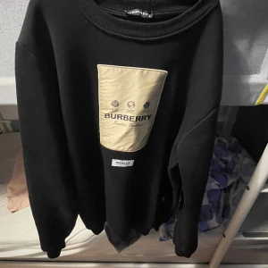 Svart Burberry sweatshirt med beige detalj - Svart sweatshirt från Burberry med stor beige patch och logga framtill. Tröjan har rund halsringning och långa ärmar. Snygg och stilren design med klassisk Burberry-känsla, perfekt för dig som gillar exklusiva streetwear-plagg. Det är storlek L men passar i M