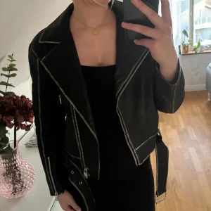 Svart skinnjacka från Nelly med bälte - Säljer en svart skinnjacka från Nelly med markerade sömmar, silvriga dragkedjor och bälte i midjan. Jackan har klassisk bikerstil med bred krage och detaljerade kanter🖤aldrig använd, skriv vid frågor
