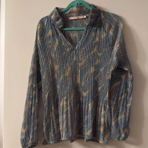 Mönstrad blus från Rue de Femme XL - Säljer en luftig blus från Rue de Femme i storlek XL. Skjortan har ett blått och beige paisleymönster med inslag av rosa, v-ringning och långa ärmar med resår vid handlederna. Materialet är lätt och känns som polyester, perfekt för dig som gillar unika prints.