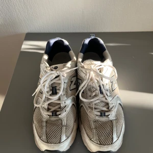 Vita New Balance sneakers med mesh - Säljer ett par klassiska New Balance sneakers i vitt och grått med marinblå detaljer. Skorna har ventilerande meshpaneler, snörning och en bekväm platt sula. Perfekta för dig som gillar sportig och retro stil. Använda ca 8 månader men är i bra skicka behövs endast tvättas upp lite. 