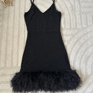 Svart kort klänning med fjädrar - Supersnygg svart kort klänning med tunna axelband, små glittriga detaljer över hela tyget och en fluffig fjäderkant längst ner. Perfekt för dig som vill sticka ut och känna dig extra glam på nästa fest! Sitter figurnära och har en modern look.