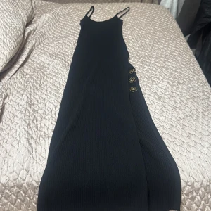 Svart långklänning från Bershka - Supersnygg svart långklänning från Bershka med smala axelband och ribbad struktur. Klänningen har dekorativa guldfärgade knappar på sidan som ger en extra touch. Perfekt för dig som gillar stilrena och trendiga plagg. Det är storlek m men den är väldigt liten men stretchiga jag har xs och den sitter bra på mig och även min mamma som har m 