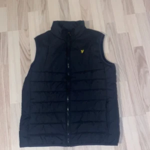 Svart dunväst från Lyle & Scott - Snygg svart dunväst från Lyle & Scott med dragkedja framtill. Västen har en quiltad design och en liten gul logga på bröstet. Perfekt för kyligare dagar.