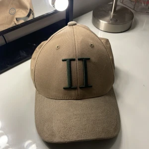 Beige keps från Les Deux - Snygg beige keps från Les Deux med broderad grön II-logga framtill och små broderade detaljer i svart, vitt och rött på sidan. Kepsen är tillverkad i en mix av bomull och polyester och har klassisk böjd skärm och justerbar baksida.