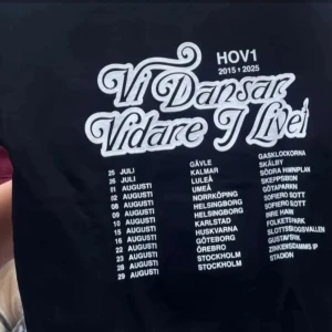 Svart Hov1 hoodie med turnétryck - Svart hoodie från Hov1 med vit kronlogga framtill och stort turnétryck på ryggen med texten 'Vi Dansar Vidare I Livet'. Hoodien har huva och magficka. Säljer helt ny pga har inte kommit till användning 