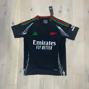 Arsenal svart matchtröja Adidas - Svart Arsenal fotbollströja från Adidas med gröna och röda detaljer på axlarna, klubbmärke i rött och vit Emirates-sponsor på bröstet. Kortärmad modell i lätt funktionsmaterial, perfekt för träning eller match.
