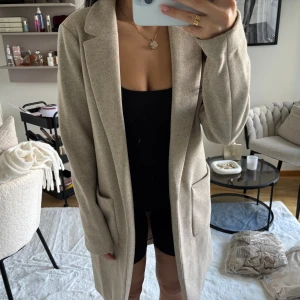 Beige kappa med stora fickor - Stilren beige kappa med klassisk krage och två stora fickor framtill. Jackan har rak passform och är helfodrad, perfekt för dig som gillar en clean och tidlös look. Materialet känns mjukt och ger en exklusiv känsla. Passar perfekt till lager-på-lager under höst och vår.