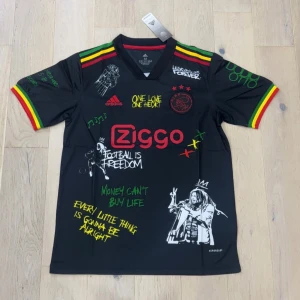 Ajax x Bob Marley svart fotbollströja - Unik svart Ajax fotbollströja från Adidas med Bob Marley-tema, Ziggo-logga och graffiti-inspirerade texter. Färgglada ränder i rött, gult och grönt på axlar och ärmslut. Tryck med musiknoter, citat och Bob Marley-illustration både fram och bak. Tillverkad i lätt polyester.