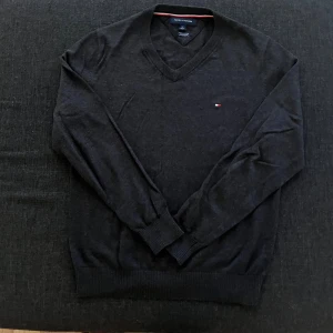 Svart v-ringad tröja Tommy Hilfiger - Svart v-ringad tröja från Tommy Hilfiger med diskret logga på bröstet. Tröjan har långa ärmar och ribbade muddar vid ärmslut och nederkant. Perfekt för dig som gillar stilrena och klassiska plagg.