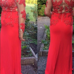 Röd långklänning med spets och pärlor - Snygg röd långklänning med transparenta ärmar och rygg, dekorerad med röda spetsapplikationer och pärlor. Klänningen har figurnära passform, lång ärm och dragkedja i ryggen. Perfekt för bal eller festliga tillfällen.