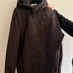 Svart vindjacka från McKinley M - Svart vindjacka från McKinley i storlek medium. Jackan har huva, hel dragkedja framtill och är tillverkad i ett lätt syntetmaterial. Perfekt för blåsiga dagar och enkel att matcha med olika outfits.