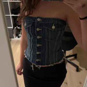 Jeans korsett tubtopp Zara - Säljer en snygg jeans korsett tubtopp med fransig nederkant och silverfärgade knappar framtill. Toppen är axelbandslös och har dekorativa fickor upptill. Perfekt för dig som gillar en edgy och trendig stil.