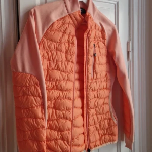 Quiltad orange jacka Peak Performance - Säljer en snygg quiltad hybrid jacka med dun från Peak Performance i orange och ljusrosa. Jackan har dragkedja framtill, bröstficka med zip och långärmade paneler i mjukt material. Perfekt för dig som gillar sportig och fräsch stil.