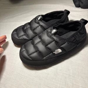 Svarta tofflor från The North Face - Mysiga svarta tofflor från The North Face med quiltad ovandel och platt sula. Perfekta att glida runt i hemma eller på fjällstugan. Tillverkade i syntetmaterial med mjuk insida och praktisk hälflik för enkel påtagning. Logga på ovansidan.