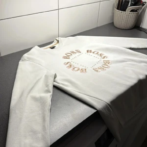 Vit sweatshirt från Hugo Boss - Vit sweatshirt från Hugo Boss med rund hals och tryck i guld och beige på bröstet där det står 'BOSS' i cirkel. Tröjan har långa ärmar och är tillverkad i mjuk bomull. Perfekt för en clean och stilren look.