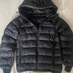 Svart dunjacka Peak Performance - Säljer en svart dunjacka från Peak Performance med huva och dragkedja. Jackan är quiltad med breda kanaler, har två fickor med dragkedja och ribbade muddar vid ärmsluten. Perfekt för kalla dagar och riktigt snygg passform.