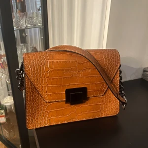Brun axelväska från Christian Laurier - Snygg brun axelväska från Christian Laurier Paris i croco-präglat skinn. Väskan har en stilren fyrkantig form, svart metallspänne och kedjedetaljer på axelremmen. Perfekt för dig som vill ha en trendig och elegant accessoar.