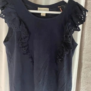 Mörkblå ärmlös topp Holly & Whyte XS - Supersöt mörkblå ärmlös topp från Holly & Whyte med volangdetaljer och broderade spetsar längs axlarna. Perfekt för dig som gillar feminina detaljer och enkel stil. Tillverkad i mjuk bomull som känns skön mot huden.