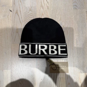 Svart mössa från Burberry - Snygg svart mössa från Burberry med vit, bred kant där det står BURBERRY i stora bokstäver runtom. Klassisk beanie-modell i mjukt stickat material, perfekt för kalla dagar. Stilsäker accessoar med tydlig logga och exklusiv känsla.