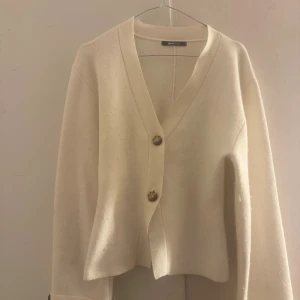 Vit kofta från Gina Tricot med knappar - Superfin vit kofta från Gina Tricot med två stora dekorativa knappar framtill. Koftan har v-ringning, lång ärm och är gjord i ett mjukt, stickat material. Perfekt för lager-på-lager och enkel att styla till olika outfits. Strl s och använd ett få tal gånger💗