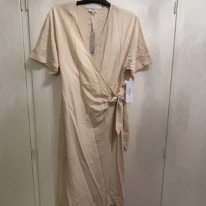 Beige omlottklänning från Happy Holly - Snygg beige omlottklänning från Happy Holly med korta ärmar och knyt i midjan. Klänningen har v-ringning och är tillverkad i ett mjukt bomullstyg. Perfekt för dig som gillar en enkel men trendig look.