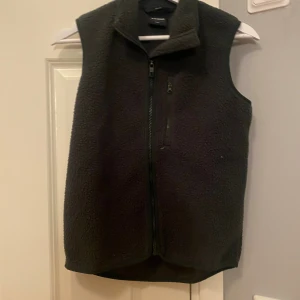 Svart fleeceväst från Everest - Mysig svart fleeceväst från Everest med hög krage och hel dragkedja framtill. Västen har en bröstficka med dragkedja och är perfekt för lager-på-lager-stil. Enkel och stilren design som funkar till det mesta.